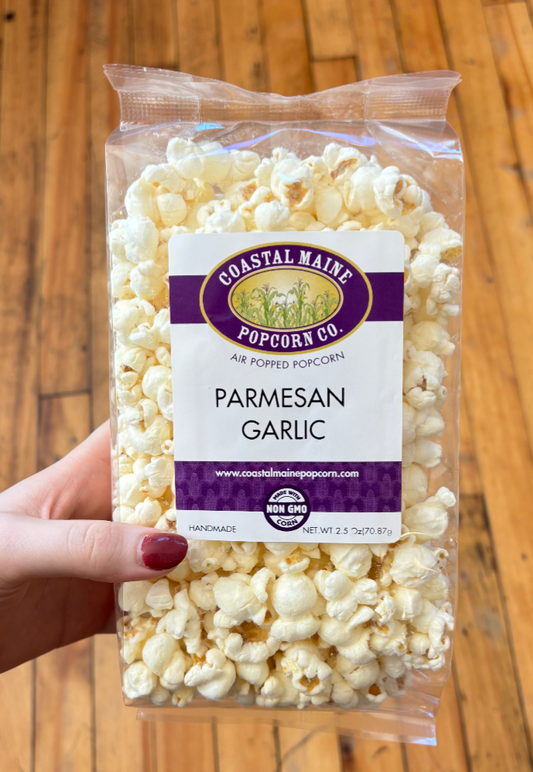 A hand holds a clear plastic bag of Coastal Maine Popcorn Co. Parmesan Garlic popcorn against a wooden floor background.